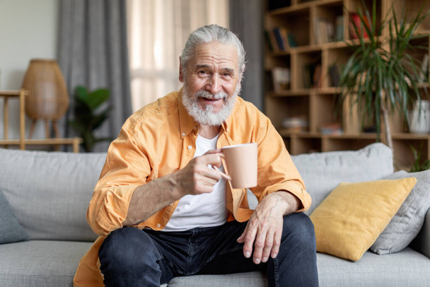 Pohledný šedovlasý starší muž pije kávu a usmívá se, sedí na gauči v pohodlném obývacím pokoji, drží šálek bylinkového čaje, usmívá se do kamery, chladí doma, kopíruje prostor - Fotografie, Obrázek