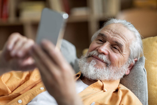 Uvolněný usměvavý starší muž ležící doma na gauči, používající moderní mobilní telefon, surfující na internetu, zatímco odpočívající o víkendu sám, detailní záběr. Volný čas a gadgets, senioři životní styl - Fotografie, Obrázek