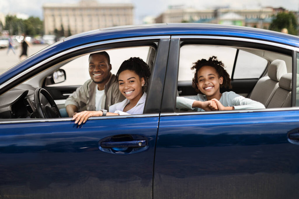 Feliz familia negra en nuevo coche azul posando sonriendo a la cámara teniendo paseo en la ciudad. Padres e hijas preadolescentes sentados en un auto de lujo. Concepto de propiedad y compra de automóviles. Enfoque selectivo - Foto, Imagen