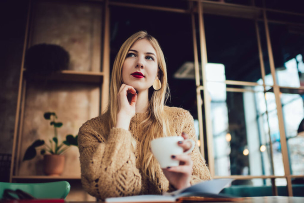 Low angle of thoughtful female in casual wear with red lipstick looking away while drinking cup of hot coffee and spending time in cafe - 写真・画像