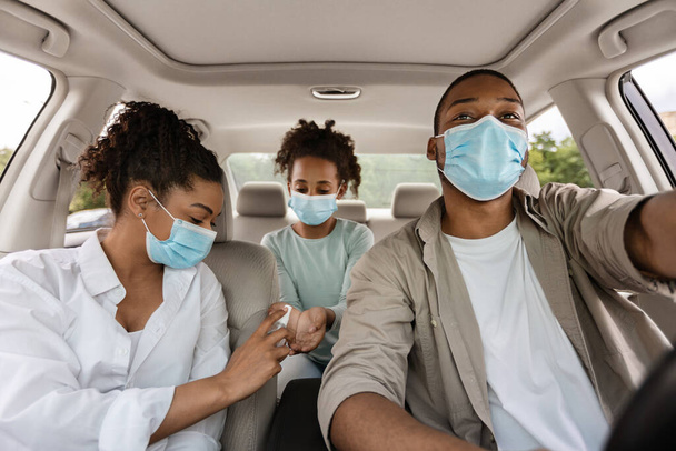 Siyah aile dezenfektan kullanıyor. Koruyucu yüz maskeleri takıyor. Arabayla seyahat ediyor. Covid-19 Salgın Salgını sırasında Güvenli Yol Gezisi. Seçici Ebeveynlere Odaklanma - Fotoğraf, Görsel