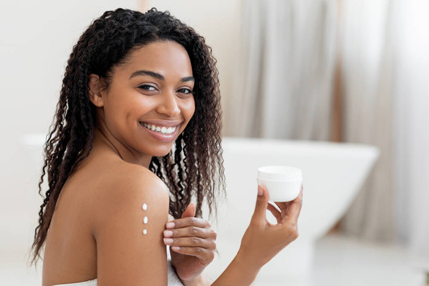 Selbstversorgungskonzept. Junge schwarze Frau, die feuchtigkeitsspendende Körperlotion auf die Haut aufträgt, glückliche Afroamerikanerin mit drei Punkten Pflegecreme auf der Schulter posiert im Badezimmerinnenraum, Kopierraum - Foto, Bild