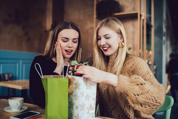 Excited female friends in casual clothes smiling and exchanging presents while sitting at wooden table in cozy modern cafe - Фото, изображение
