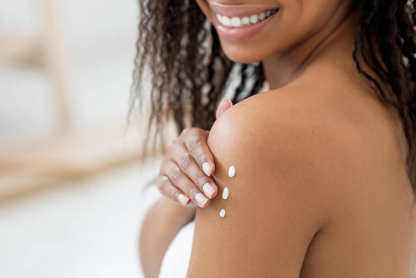 Mausernde Haut. Lächelnde schwarze Frau, die nährende Körperlotion aufträgt, Nahaufnahme einer unkenntlichen jungen Afroamerikanerin, die nach dem Duschen eine Selbstpflege-Routine durchführt, beschnittenes Bild - Foto, Bild