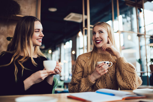 Optimistic female friends in casual wear sitting at wooden table with cup of coffee and talking to each other in cafe - Foto, Bild