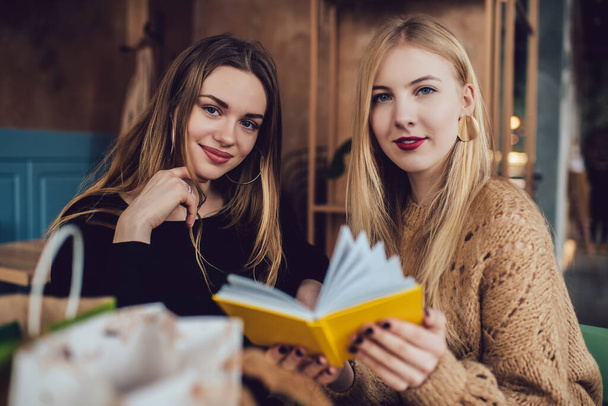 Young charming cheerful women holding yellow copybook and looking at camera with bright toothy smile while spending time together in cafe - Фото, зображення