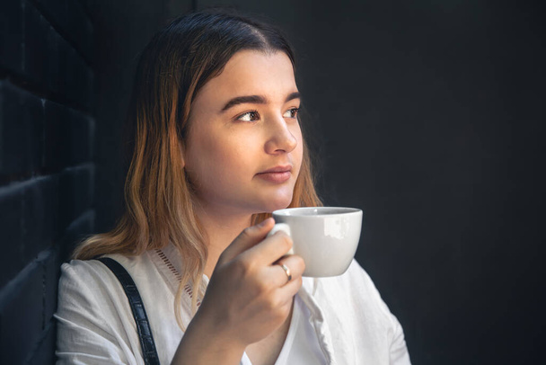Μια νεαρή γυναίκα με ένα φλιτζάνι καφέ σε ένα μαύρο εσωτερικό καφέ απολαμβάνει ένα ποτό, αντίγραφο χώρο. - Φωτογραφία, εικόνα