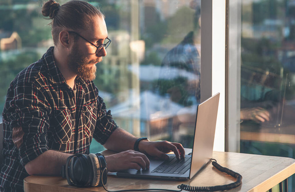 Un homme avec une barbe travaille à un ordinateur, assis près d'une fenêtre donnant sur la ville, le concept d'informatique, freelance et travail à distance. - Photo, image