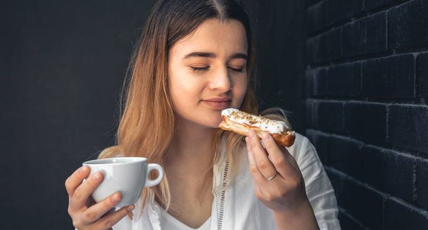 A young woman enjoys a cup of coffee and an eclair on a black background of a cafe interior. - Foto, Imagem