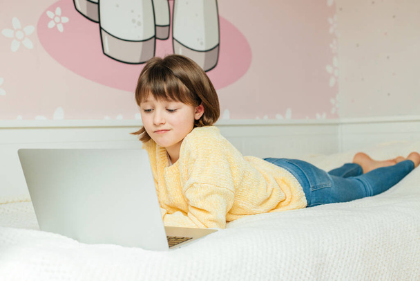 Afstandsonderwijs online thuis. Een schoolmeisje ligt op een bed met een laptop in haar kamer. Kinderen communiceren online - Foto, afbeelding