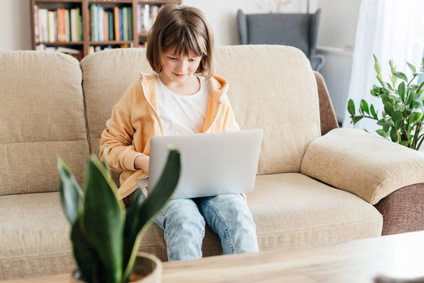 Hausaufgabenbetreuung für Kinder. Ein Mädchen sitzt mit einem Laptop auf dem Schoß auf einem Sofa und schaut sich Webinare oder Chats mit Freunden online an. Kinder nutzen Technik - Foto, Bild