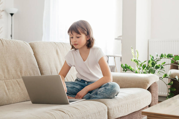 The schoolgirl sits on the couch and writes in a laptop. The child is learning using gadgets at home or chatting with friends. Children use technology in everyday life - 写真・画像