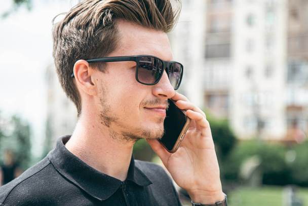 Güneş gözlüklü çekici genç adam yazın şehrin arka planında akıllı bir telefonla konuşuyor.. - Fotoğraf, Görsel