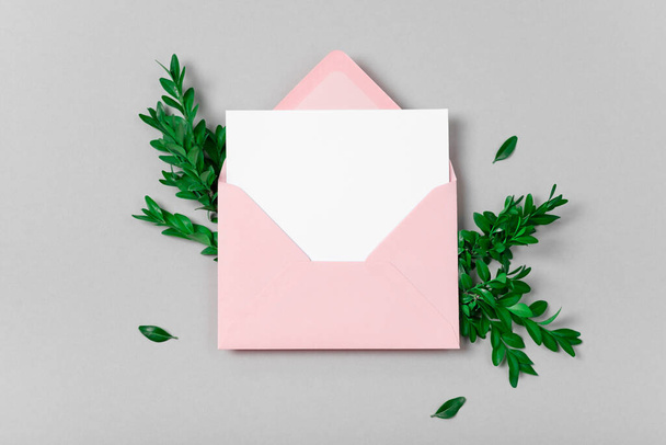 Real photo. Pink envelope square invitation white greeting card mockup with a boxwood branch. Top view with copy space, pastel grey background. Template for branding and advertising - 写真・画像