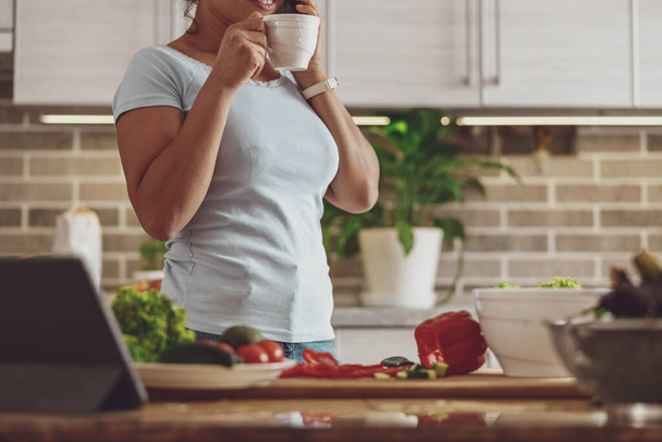 Gospodyni domowa pauzy w przygotowaniu sałatki warzywnej popijając herbatę stojącą w kuchni - Zdjęcie, obraz