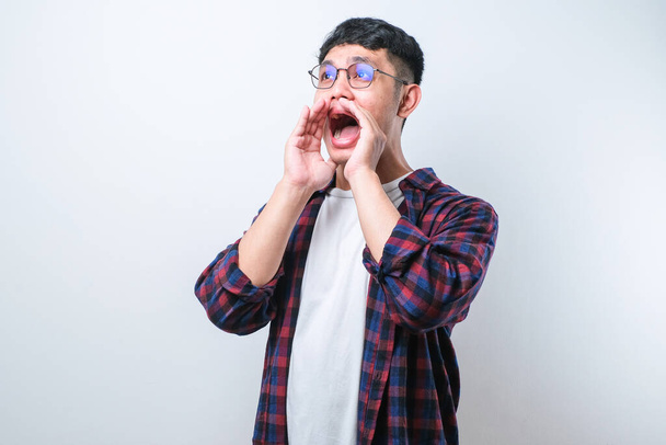 Junger asiatischer Mann in lässigem Hemdstil schreit und schreit laut mit der Hand auf dem Mund vor weißem Hintergrund. Kommunikationskonzept. - Foto, Bild