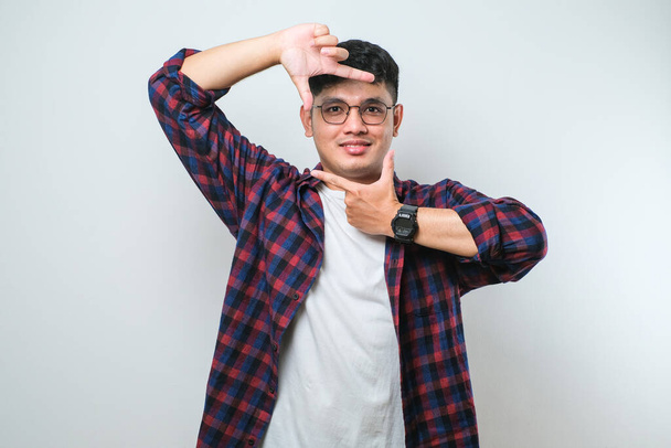Young handsome asian boy wearing casual shirt standing over isolated white background smiling making frame with hands and fingers with happy face. Creativity and photography concept - Zdjęcie, obraz