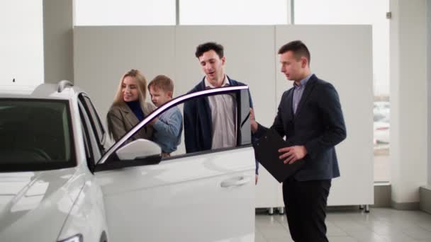 νεαρός άνδρας και γυναίκα με το μικρό αγόρι στην αγκαλιά τους επιλέγουν νέο αυτοκίνητο για την οικογένειά τους, μιλώντας με τον αρσενικό πωλητή στο κέντρο πωλήσεων αυτοκινήτων - Πλάνα, βίντεο