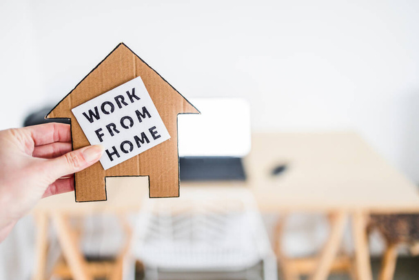 work from home sign being hold in front of out of focus home office desk setup, concept of digital nomads working remotely or wfh days during lockdowns or covid-19 isolation - Foto, Imagem