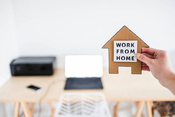 work from home sign being hold in front of out of focus home office desk setup, concept of digital nomads working remotely or wfh days during lockdowns or covid-19 isolation - Foto, Imagen