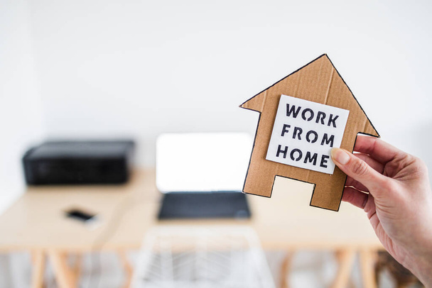 work from home sign being hold in front of out of focus home office desk setup, concept of digital nomads working remotely or wfh days during lockdowns or covid-19 isolation - 写真・画像