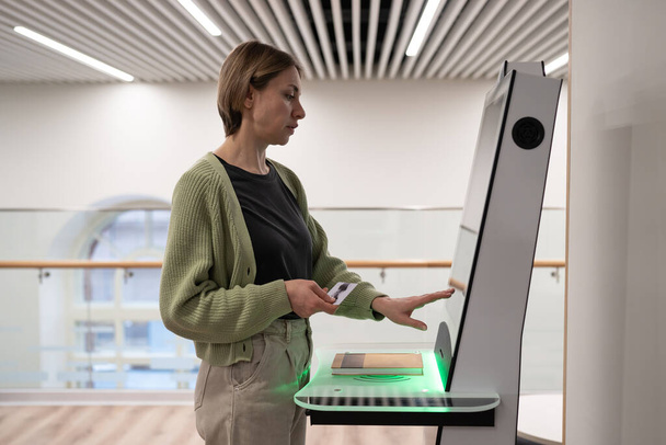 Middle-aged woman using self-service terminal in digital library space, registering book, searching and selecting literature or browsing catalogue. Innovative technologies in libraries - Zdjęcie, obraz