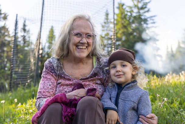 Primer plano retrato alegre de anciana abuela sonriente con el brazo alrededor lindo nieto de cuatro años sentado al aire libre en la naturaleza con fondo borroso. - Foto, Imagen