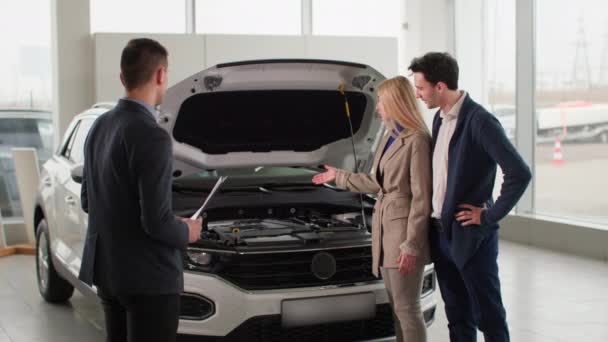 car business, professional manager dealership employee advises young couple on choosing new automobile model in a auto center - Materiał filmowy, wideo