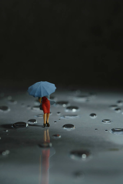 Persone in miniatura giocattolo figura fotografia. Una donna che indossa un impermeabile con l'ombrello, che cammina per strada durante una tempesta, contro un vento molto forte. Nuvoloso scuro, sfondo temporale. Foto immagine - Foto, immagini
