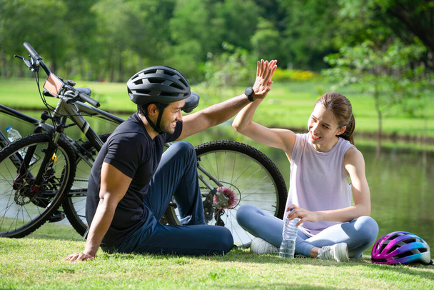 jong aziatisch paar handen hoog vijf samen terwijl rusten naast de vijver in het park, gelukkig gezin zitten in de zon na rit fiets in de ochtend. - Foto, afbeelding