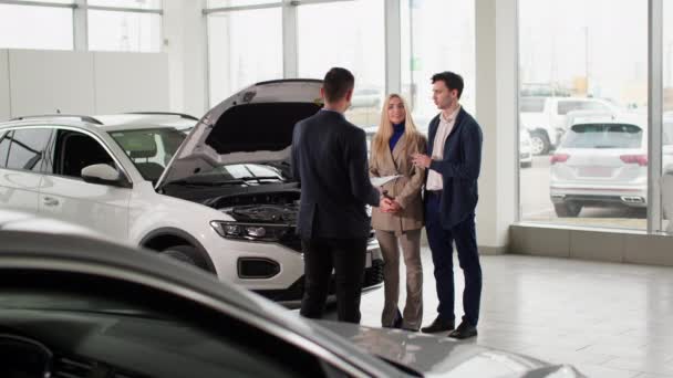 дилерство, молодой человек и женщина консультируют менеджера автосалона по новой модели автомобиля в центре продаж автомобилей - Кадры, видео