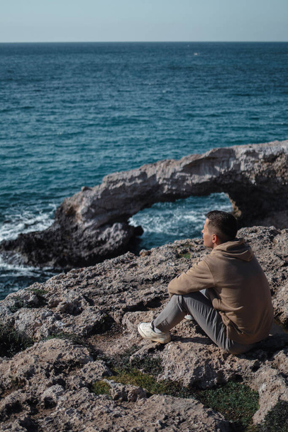 A young man from behind is sitting alone at the edge and looking at the sea cave arch bridge of love in Ayia Napa, Cyprus - 写真・画像
