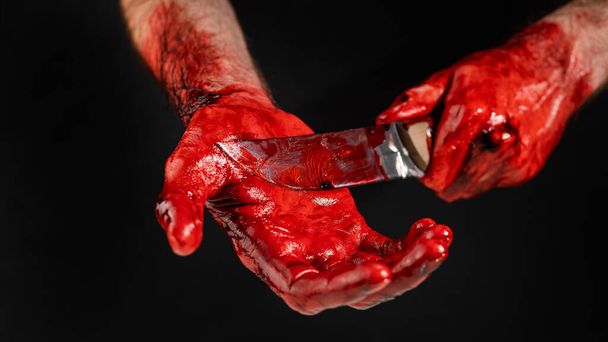 A man with blood-stained hands cuts his palm with a knife on a black background - 写真・画像