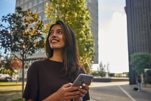 Young happy businesswoman standing in sun smiling and holding mobile phone - Photo, image