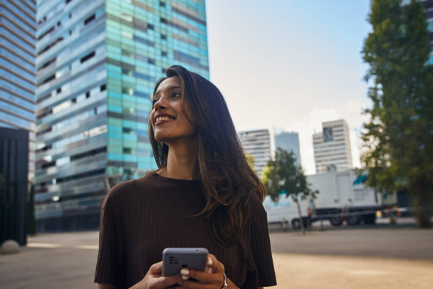 Happy cute businesswoman holding mobile phone when standing on street against blue sky - Foto, Bild