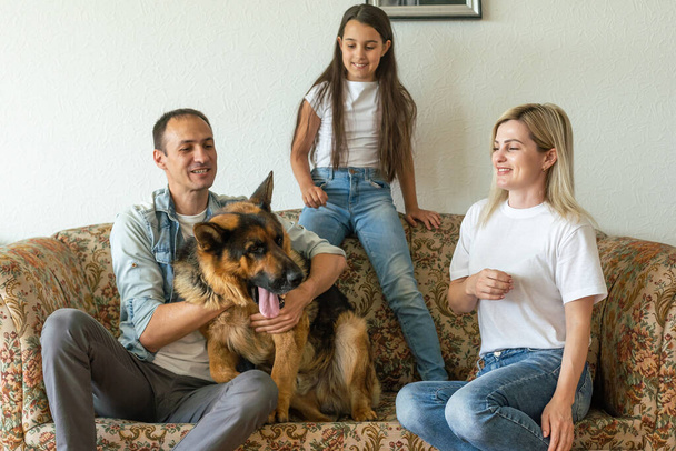 Beaux jeunes parents, leur jolie petite fille regardant la caméra et souriant, assis avec leur chien mignon sur le canapé à la maison. - Photo, image