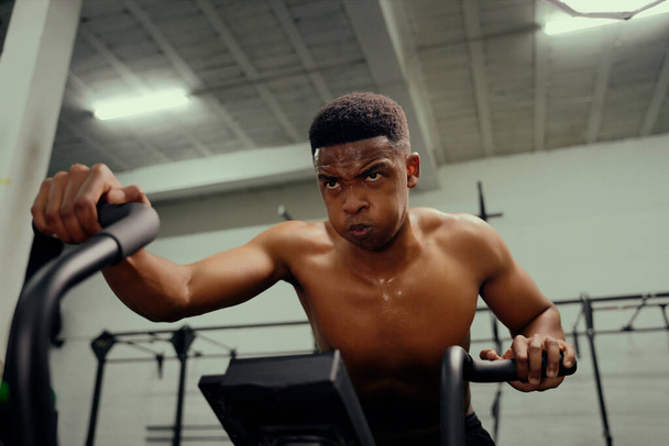 African American male using an elliptical trainer during cross fit training. Male personal trainer exercising intensely in the gym. High quality photo - 写真・画像