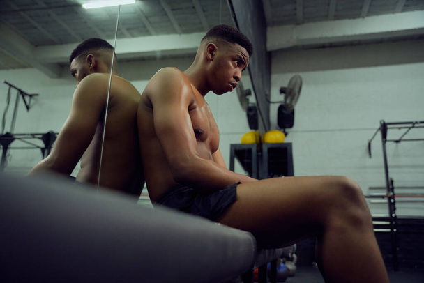 African American male athlete resting in the gym after a hard cross training. Shirtless mixed race male sitting down after exercising. High quality photo - Zdjęcie, obraz