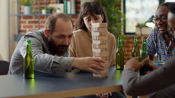 Positive people having fun with society game in living room, playing with wooden tower blocks. Men and women enjoying game with building square pieces on structure for entertainment. - Photo, Image