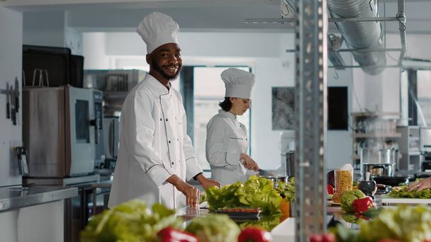 Authentic portrait of african american cook preparing food dish for restaurant menu in professional kitchen. Male chef cooking gastronomy gourmet meal with recipe ingredients, cookery service. - 写真・画像
