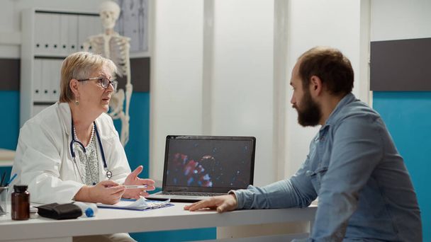 Female medic looking at coronavirus illustration on laptop with sick patient, talking about medication and prevention. Virus animation on display to cure disease in medical cabinet. - Photo, Image