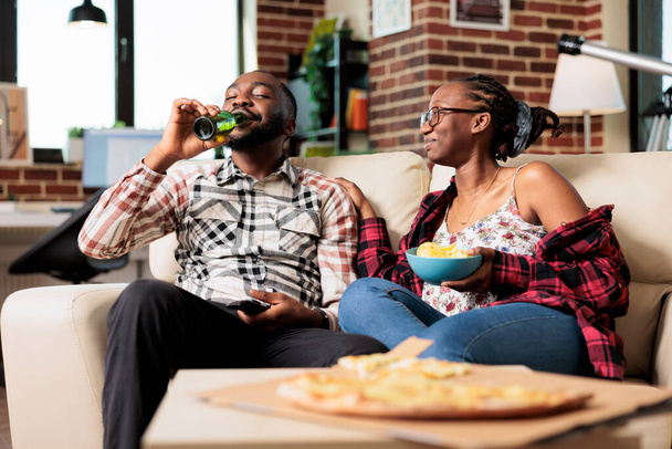 Африканские американцы веселятся с закусками и фильмами по телевизору, переключая программу канала с дистанционным управлением. Есть чипсы и еду на вынос, смотреть фильм по телевизору. - Фото, изображение