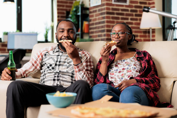 African american couple eating slice of pizza and drinking bottles of beer while they watch movie on tv together. Enjoying fast food takeaway delivery and beverage, watching television. - Фото, изображение