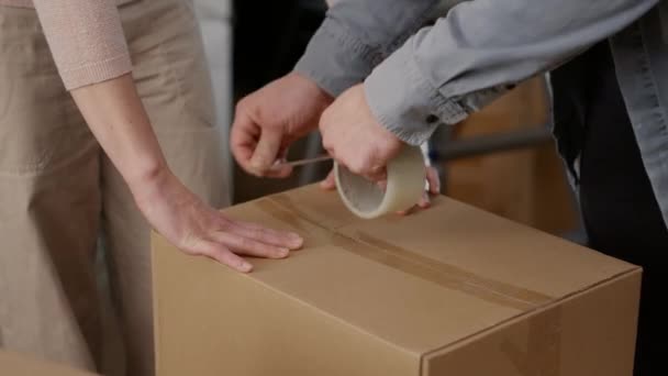 Couple using adhesive tape to pack things in cardboard boxes, wrapping carton storage containers with sticky roller. Preparing packages for shipping transportation to move in. Close up. - Filmmaterial, Video