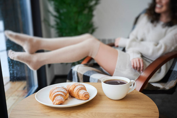 Místo s croissanty a šálkem čaje na stole. Oříznutý pohled na mladé ženy nohy v blízkosti okna uvnitř současného stodoly dům na rozmazaném pozadí. - Fotografie, Obrázek