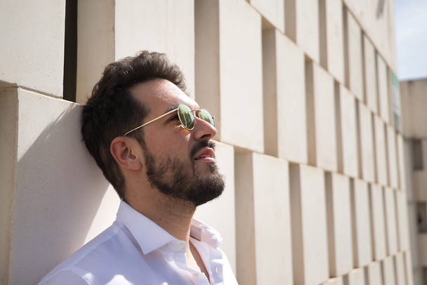 Portrait of handsome young man with beard, sunglasses and white shirt with his head leaning against a white stone block wall. Concept beauty, fashion, trend, sun, loneliness. - Photo, image