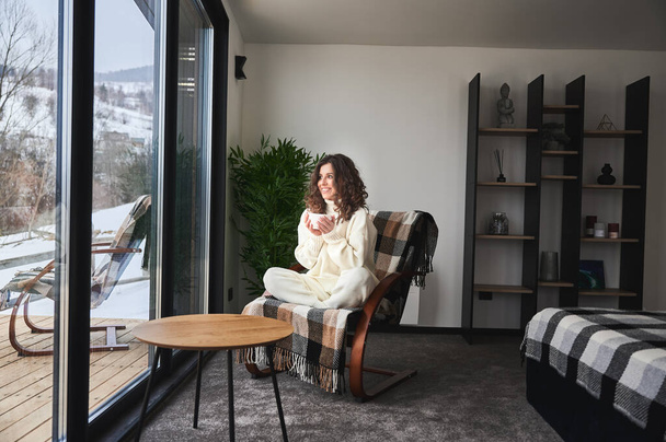 Curly young woman resting inside contemporary barn house in the mountains. Happy female tourist sitting on chair, holding cup tea, enjoying view of mountain landscape through panoramic windows. - Foto, imagen
