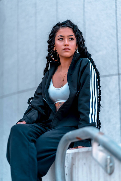 Urban session. Young woman of black ethnicity with long braids and with tattoos, sitting in the city, vertical photo - Foto, Imagem