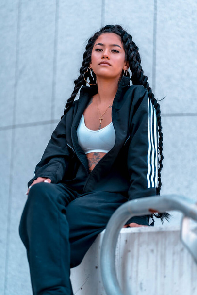 Urban session. Young woman of black ethnicity with long braids and with tattoos, sitting in the city, vertical photo - Photo, Image