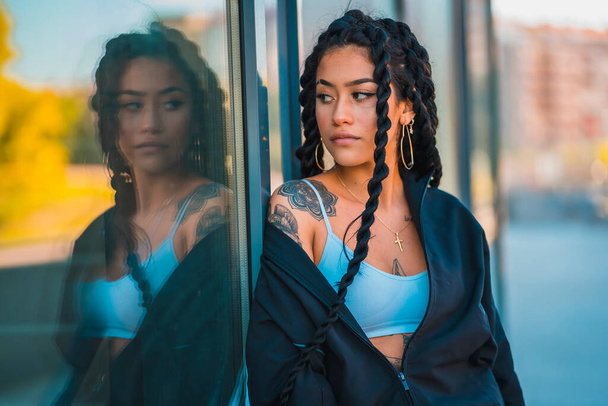 Urban session. Young woman of black ethnicity with long braids and with tattoos, reflected in glass - Photo, Image